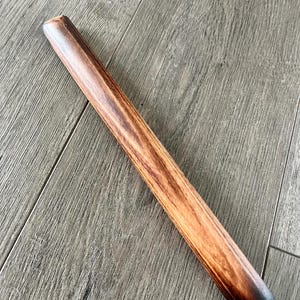 May include: A wooden rolling pin with a dark brown finish and a rounded, tapered end. The wood grain is visible, and the pin is lying on a wooden surface. The rolling pin is a kitchen tool used for baking.