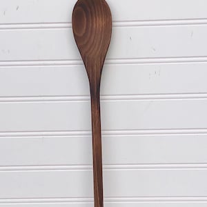 May include: A long, wooden spoon with a dark brown stain. The spoon has a rounded, oval-shaped head.