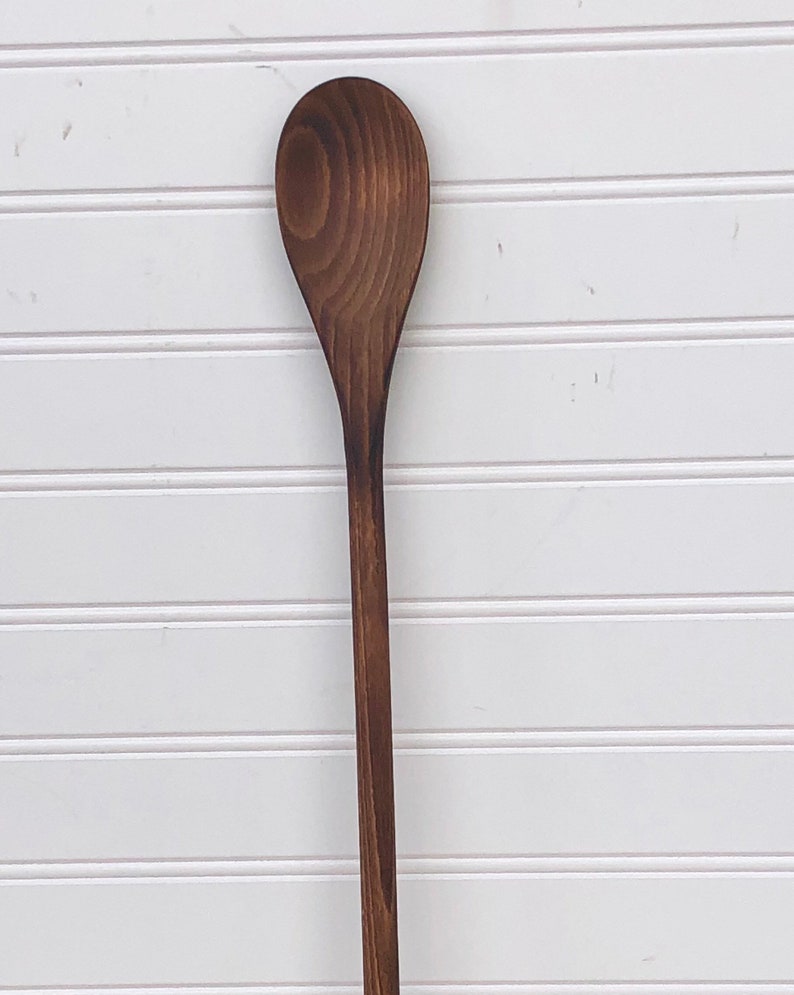 May include: A long, wooden spoon with a dark brown stain. The spoon has a rounded, oval-shaped head.