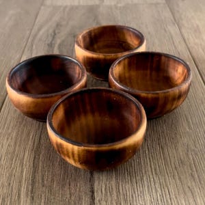May include: Four small wooden bowls with a dark brown, almost black, finish. The bowls are arranged in a semi-circle on a wooden surface.
