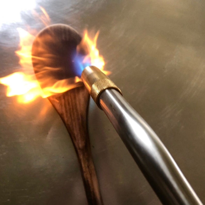 May include: A wooden spoon is being torched with a blue and orange flame from a handheld blowtorch. The blowtorch is made of metal with a brass tip.