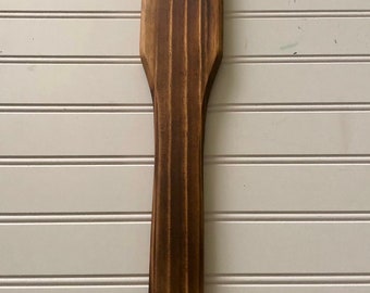 May include: A wooden spatula with a dark brown stain. The spatula has a wide, flat head and a long, narrow handle.