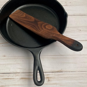 May include: A black cast iron skillet with a wooden spatula resting inside. The skillet has a loop handle and is sitting on a white wooden surface.