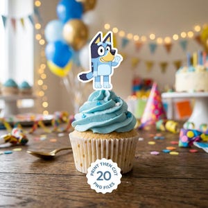Op de afbeelding: Een cupcake topper met een blauwe tekenfilmhond met een crèmekleurige snuit en buik, staand met één arm omhoog. De topper staat op een lichtblauw geglazuurde cupcake met een wit papieren vormpje en een rond label eronder.