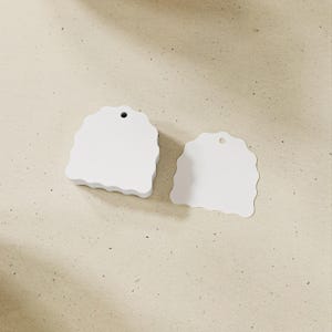 May include: Two white, arch-shaped tags with wavy, scalloped edges. Each tag features a small, circular hole at the top center for hanging. The larger tag on the left has a thicker profile, while the smaller tag on the right is flat.
