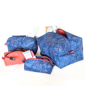 May include: A collection of travel cosmetic bags in various sizes. The bags are primarily blue with a swirling pattern and red accents. One bag is coral-colored. The bags are open, revealing toiletries and a USB cable.