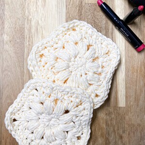 Pode incluir: Duas toalhas de croch&ecirc; quadradas creme com um design floral. As toalhas s&atilde;o feitas de um fio macio e texturizado. O fundo inclui itens de maquiagem em uma superf&iacute;cie de madeira.