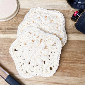 Pode incluir: Tr&ecirc;s esponjas faciais quadradas de croch&ecirc; creme com um design floral s&atilde;o dispostas sobre uma superf&iacute;cie de madeira. As esponjas feitas &agrave; m&atilde;o s&atilde;o feitas de um fio macio e texturizado. Uma embalagem de maquiagem e uma bolsa de cosm&eacute;ticos preta s&atilde;o vis&iacute;veis no fundo.