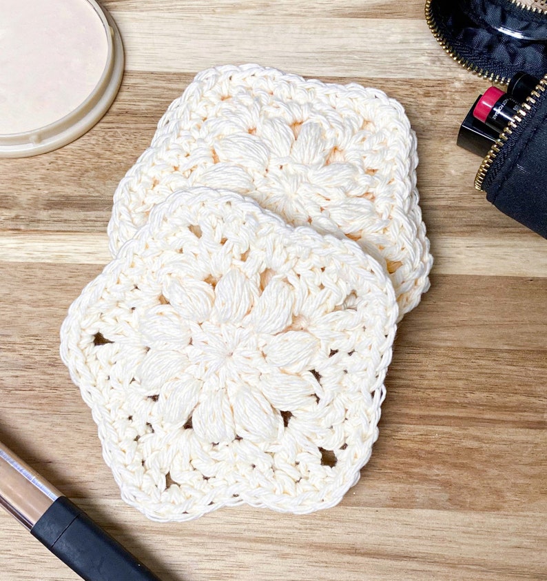 Pode incluir: Tr&ecirc;s esponjas faciais quadradas de croch&ecirc; creme com um design floral s&atilde;o dispostas sobre uma superf&iacute;cie de madeira. As esponjas feitas &agrave; m&atilde;o s&atilde;o feitas de um fio macio e texturizado. Uma embalagem de maquiagem e uma bolsa de cosm&eacute;ticos preta s&atilde;o vis&iacute;veis no fundo.