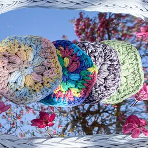 Pode incluir: Quatro itens circulares de croch&ecirc; coloridos em tons pastel de amarelo, azul, verde e roxo. Os itens est&atilde;o dispostos em uma fileira, refletindo em um espelho com flores rosa e um fundo de c&eacute;u azul.