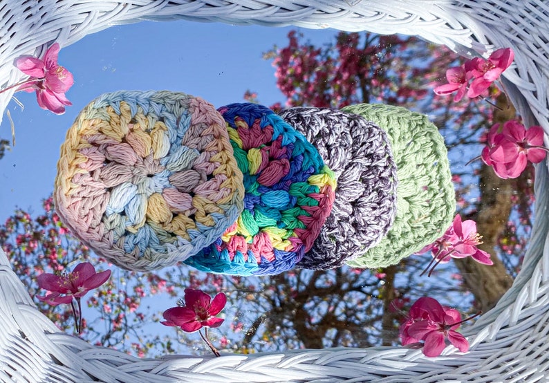 Pode incluir: Quatro itens circulares de croch&ecirc; coloridos em tons pastel de amarelo, azul, verde e roxo. Os itens est&atilde;o dispostos em uma fileira, refletindo em um espelho com flores rosa e um fundo de c&eacute;u azul.