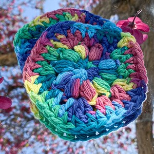 Pode incluir: Dois itens redondos e coloridos, feitos de croch&ecirc; em tons de azul, verde, rosa e amarelo. Os itens s&atilde;o empilhados uns sobre os outros. O fundo &eacute; uma vis&atilde;o desfocada de uma &aacute;rvore com flores rosas.