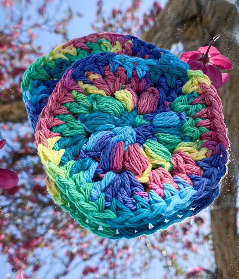 Pode incluir: Dois itens redondos e coloridos, feitos de croch&ecirc; em tons de azul, verde, rosa e amarelo. Os itens s&atilde;o empilhados uns sobre os outros. O fundo &eacute; uma vis&atilde;o desfocada de uma &aacute;rvore com flores rosas.