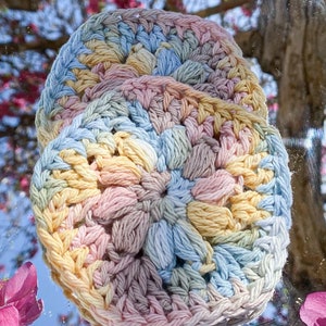 Pode incluir: Duas bases para copos redondas, feitas &agrave; m&atilde;o em croch&ecirc;, em cores pastel. As bases apresentam um padr&atilde;o texturizado em relevo em tons de amarelo, azul, rosa e bege. As bases s&atilde;o exibidas em um fundo desfocado de flores rosas e uma &aacute;rvore.