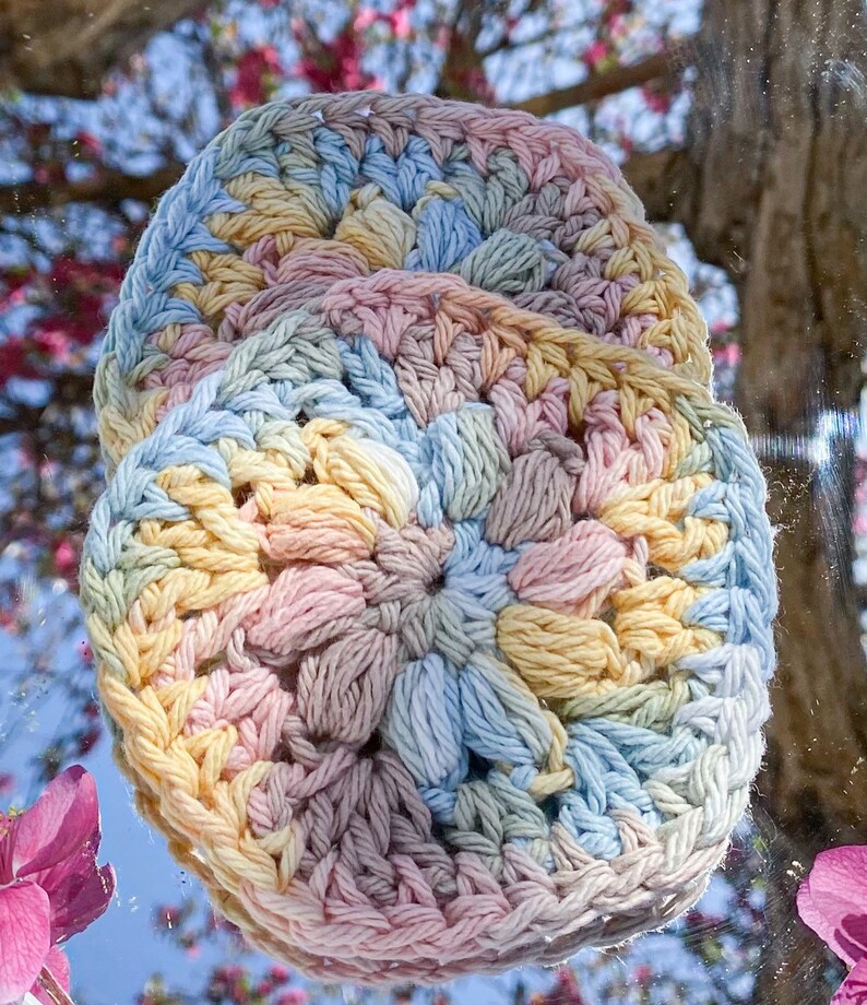 Pode incluir: Duas bases para copos redondas, feitas &agrave; m&atilde;o em croch&ecirc;, em cores pastel. As bases apresentam um padr&atilde;o texturizado em relevo em tons de amarelo, azul, rosa e bege. As bases s&atilde;o exibidas em um fundo desfocado de flores rosas e uma &aacute;rvore.