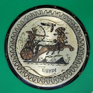 May include: A round, silver-colored metal plate with a black border and intricate gold designs. The center features a scene of a chariot pulled by a horse, with a person riding in the chariot. The text "Hurghada Egypt" is printed below the scene.