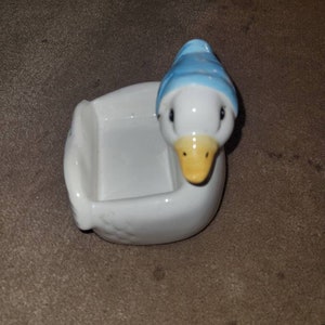 May include: White ceramic goose figurine with a blue hat and a yellow beak. The goose has a small dish on its back.