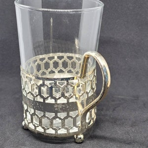 May include: A silver-tone glass holder with a clear glass cup and a handle. The holder has a geometric pattern and sits on three small feet.