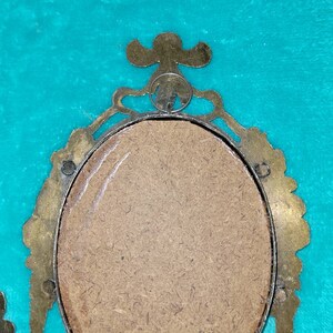 May include: A gold-toned oval picture frame with ornate detailing. The frame has a brown cardboard backing and a metal ring for hanging.