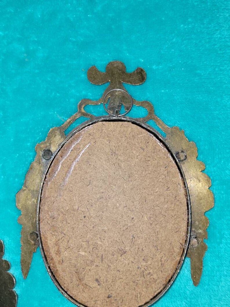 May include: A gold-toned oval picture frame with ornate detailing. The frame has a brown cardboard backing and a metal ring for hanging.