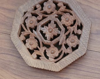 May include: A wooden, octagonal decorative piece with intricate floral carvings. The design features detailed openwork with stylized flowers and leaves. The piece is made of light brown wood and is likely for home decor.