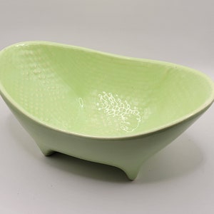 May include: A light green ceramic bowl with a textured surface and three legs. The bowl is shaped like a bathtub.
