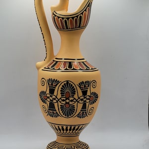 May include: A ceramic pitcher with a black and orange geometric design. The pitcher has a tall, slender body with a wide, round base. The handle is curved and has a decorative design.