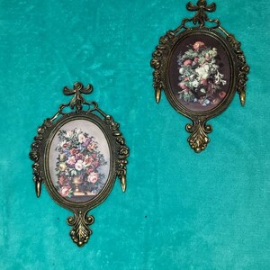 May include: Two ornate gold-toned oval picture frames with floral designs. Each frame features a vintage floral print with a vase of flowers.
