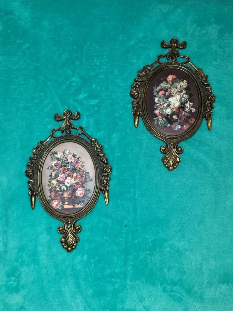 May include: Two ornate gold-toned oval picture frames with floral designs. Each frame features a vintage floral print with a vase of flowers.