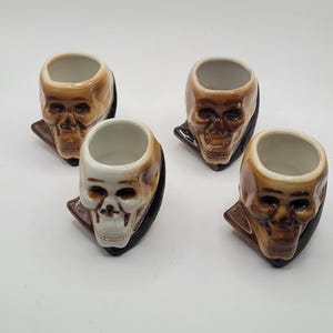 May include: Set of four ceramic shot glasses shaped like skulls with brown and white paint. Each skull has a black painted collar.