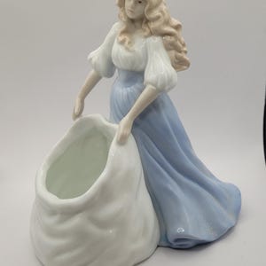 May include: A porcelain figurine of a woman in a blue dress holding a white bag. The bag is shaped like a sack and has a wide opening at the top. The figurine is a decorative piece and could be used as a planter.