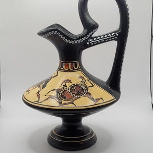 May include: Black and yellow ceramic pitcher with a black handle and a white geometric design. The pitcher features a scene of Greek warriors in black, red, and white.