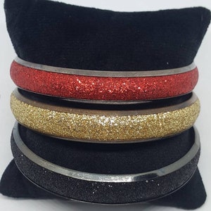 May include: Three silver bangles with glitter. The top bangle is red glitter, the middle bangle is gold glitter, and the bottom bangle is black glitter.