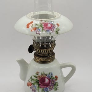 May include: A white porcelain oil lamp shaped like a teapot with a floral design. The lamp has a glass chimney and a brass burner.