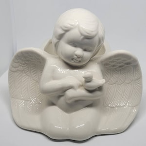 May include: White ceramic angel figurine holding a small bird. The angel is kneeling with wings outstretched and a hollow space in the center of the figurine.