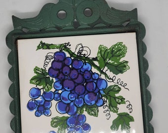 May include: Green cast iron trivet with a white ceramic tile featuring a hand-painted design of a bunch of grapes with green leaves.