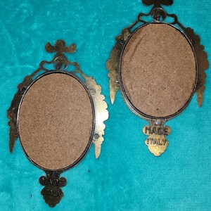 May include: Two oval-shaped brass picture frames with ornate details. The frames are empty and have a brown backing. The back of each frame has a label that reads "Made Italy".