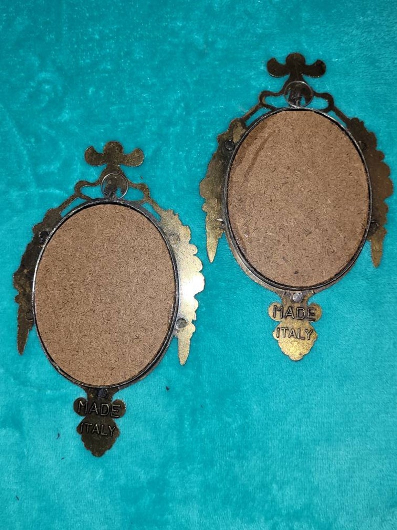 May include: Two oval-shaped brass picture frames with ornate details. The frames are empty and have a brown backing. The back of each frame has a label that reads "Made Italy".