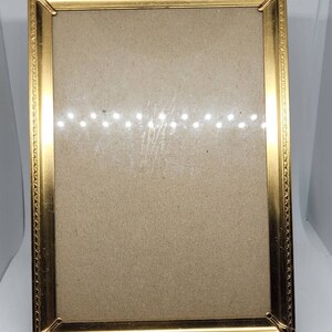 May include: A gold-colored picture frame with a simple, ornate design. The frame has a matted opening for a photo or artwork.