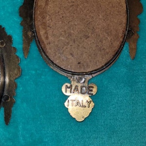 May include: A close-up of the back of a small, oval-shaped picture frame with a gold-colored metal frame. The frame has a brown backing and a gold-colored label that reads "MADE ITALY".