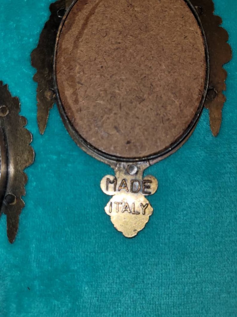 May include: A close-up of the back of a small, oval-shaped picture frame with a gold-colored metal frame. The frame has a brown backing and a gold-colored label that reads "MADE ITALY".