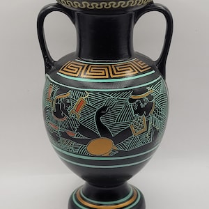 May include: A black ceramic vase with two handles, featuring a detailed illustration of figures in teal, gold, and red. The vase has a decorative rim and base with gold and teal accents, showcasing a classical design.