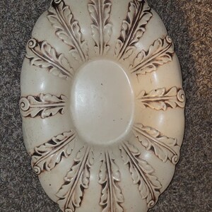 May include: A white ceramic bowl with a decorative design of brown leaves. The bowl is oval shaped and has a raised rim.