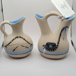 May include: Two cream-colored ceramic vases with blue rims and handles. The vases are decorated with black and turquoise Native American-inspired designs, including arrows and a pipe. The vases are of different sizes.