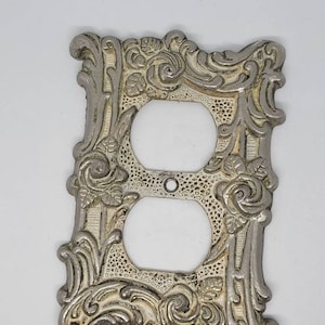 May include: Ornate silver-toned metal outlet cover with a floral design. The cover has two outlets and a decorative center piece.