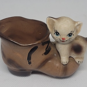 May include: A brown ceramic shoe with a white kitten sitting on top. The shoe has a black design on the side and the kitten has blue eyes and a pink nose.