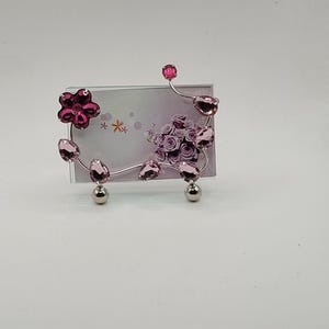 May include: A decorative photo frame with a floral design. The frame features a clear glass insert, a silver-colored metal frame, and pink crystal accents shaped like flowers and leaves. The frame is supported by two silver-colored ball feet.
