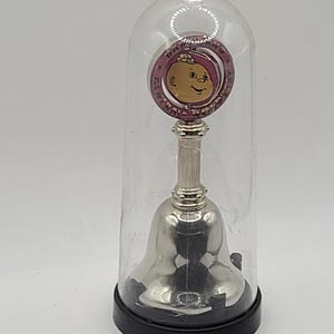 May include: A silver bell with a pink and silver charm featuring a cartoon baby face and the text "Baby's First Christmas" is displayed under a clear glass dome.