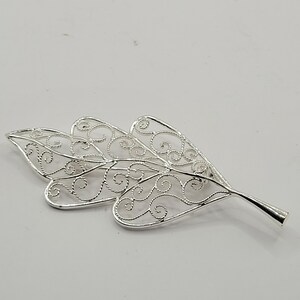 May include: A silver-colored leaf-shaped brooch with intricate openwork filigree detailing. The brooch has a stem and three main leaf sections, each with a delicate, swirling pattern. The design is symmetrical and elegant, suitable for adding a touch of nature-inspired flair.