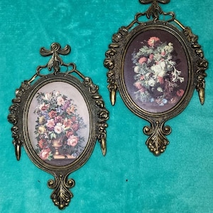 May include: Two ornate gold-toned oval frames with floral prints. Each frame has a decorative top and bottom with a floral design. The frames are hanging on a teal background.